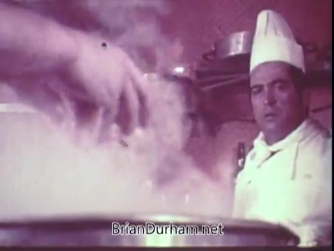 The still from the 1972 Blue Bonnet Margarine commercial features a chef in a white uniform and hat, looking intently at a bubbling pot in a kitchen. Steam rises dramatically from the pot, indicating active cooking. The scene captures the essence of culinary preparation, showcasing the use of Blue Bonnet Margarine in a visually engaging manner.