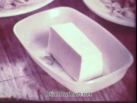 The still features a block of Blue Bonnet Margarine placed on a small, oval dish. The setting appears rustic, with a wooden table and plates of food visible in the background. The color palette is soft and slightly faded, characteristic of 16mm film from the early 1970s. The overall composition emphasizes the margarine, likely showcasing its use in cooking or as a spread.