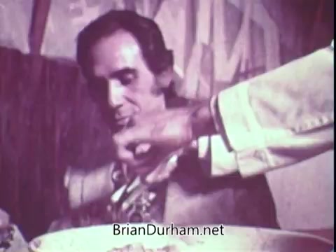 The still from the 1972 Blue Bonnet Margarine commercial features a man seated at a table, appearing content as he interacts with food. A hand, presumably of a server, gestures toward a dish, likely offering a portion. The scene has a nostalgic quality, characterized by its vintage aesthetic typical of early commercial filmmaking.