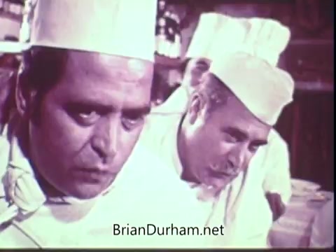 The still from the 1972 Blue Bonnet Margarine commercial features two chefs in white uniforms and hats, focused on their tasks in a kitchen setting. Their expressions convey concentration and seriousness, possibly as they prepare a dish using Blue Bonnet Margarine. The overall tone suggests a dedication to cooking and quality.