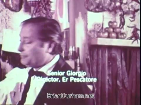 The still from the 1972 Blue Bonnet Margarine commercial features a man identified as Senior Giorgio, likely in a restaurant or culinary setting. The background includes decorative elements that suggest a warm, inviting atmosphere. The focus appears to be on the character's expressions and interactions, emphasizing a moment in the promotional narrative.