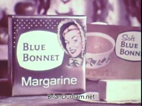 The still features packaging for Blue Bonnet Margarine, showcasing a vintage design. The iconic box displays the brand name 'Blue Bonnet' prominently, accompanied by a smiling woman wearing a bonnet. Adjacent to it is another container labeled 'Soft Blue Bonnet.' The overall aesthetic reflects the advertising style of the early 1970s, combining bright colors and a friendly, nostalgic tone.