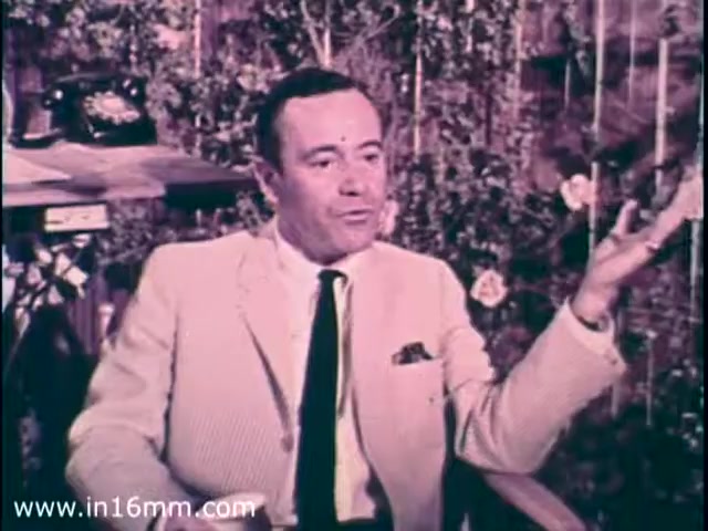 The still from the 1968 PSA features Jack Lemon seated, dressed in a light-colored suit and black tie. He appears engaged in conversation, gesturing with one hand. The background has a nature-themed setting with greenery and a telephone nearby. The overall tone is serious yet approachable, as the PSA aims to raise awareness about epilepsy.