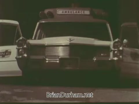 The still from the 'Aetna Life PSA - 1960s' features a prominent vintage ambulance in the foreground, displaying the word 'AMBULANCE' on its roof. Flanking the ambulance are other vehicles, including a white car on the left, suggesting a scene related to healthcare or emergency services. The overall color palette appears muted, characteristic of the era, and the composition focuses on the ambulance, highlighting its significance in the context of the public service announcement.