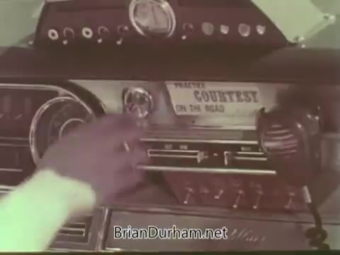The still from the Aetna Life PSA shows a close-up of a vintage car's dashboard, highlighting a radio and controls. A hand is reaching towards the radio, possibly adjusting the settings. The scene evokes a sense of nostalgia associated with driving in the 1960s, emphasizing themes of connectivity and safety on the road. Key text visible reads 'Courtesy on the Road,' hinting at the PSA's message promoting safe driving practices.