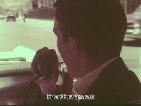 The still from the 'Aetna Life PSA - 1960s' depicts a man in a suit driving a car while speaking into a two-way radio. The scene conveys a sense of urgency or communication, set against a backdrop of a city street. The overall color tone is warm and slightly faded, characteristic of 16mm film from that era. The focus seems to be on the interaction between the driver and possibly someone on the other end of the radio, emphasizing the importance of connection and communication.