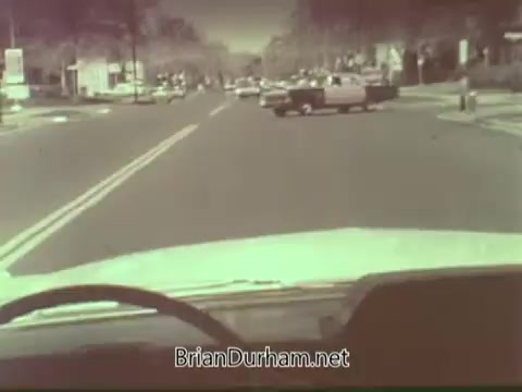 The still features a view from the perspective of a vehicle's dashboard, looking down a street during the 1960s. The scene is tinted with a vintage hue, suggesting an older film quality. Various vehicles are seen on the road, and there are trees and buildings lining the street, typical of a suburban setting. The focus is on the road ahead, creating a sense of movement and driving experience.