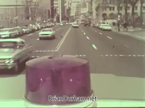The still features a view from the rooftop of a vehicle, likely an emergency or service vehicle, showcasing a busy urban street scene. Classic cars are seen in both lanes, and city buildings can be seen in the background, indicating a bustling environment of the 1960s. The prominent purple light on the vehicle suggests a focus on public safety or emergency service in the context of the Aetna Life PSA.
