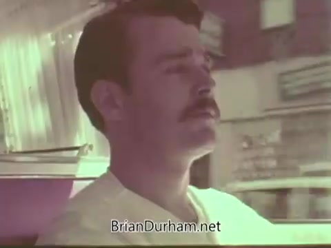 The still from the 'Aetna Life PSA - 1960's' features a man with a mustache driving a vehicle. He appears focused and contemplative, with a soft, warm color palette typical of the era. The background hints at a mid-century urban setting, likely intended to convey a message related to life insurance or personal well-being. The overall tone reflects the style of public service announcements from that time.
