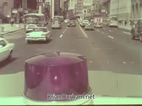 The still features a view from the front of a vehicle, showcasing a busy city street filled with various cars from the 1960s. The prominent purple light on the vehicle's roof indicates it may belong to an emergency or service vehicle. The background includes buildings typical of a bustling urban environment, reflecting the mid-20th century aesthetic. The vibrant colors and clear scene capture the dynamic traffic and energy of the era.
