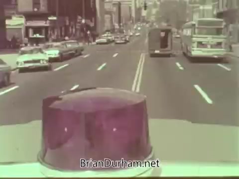 The still captures a street scene from the 1960s, viewed from the perspective of a vehicle equipped with a light on top. The street is busy with various cars and a bus, typical of urban life during that era. Buildings line the street, suggesting a city environment. The overall color tone is faded, characteristic of vintage film, likely conveying a nostalgic or historical vibe related to the Aetna Life PSA content.