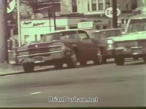 The 16mm still from the Aetna Life PSA features a busy urban street scene from the 1960s. Two cars are prominently displayed: a red vehicle on the left and a white one on the right. Background elements include storefronts and street signs, typical of the era's suburban landscape. The image captures the essence of mid-20th century American life, likely emphasizing themes of community and safety in the context of the PSA.