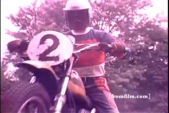 The still features a motorcyclist wearing a helmet and protective gear, sitting on a bike marked with the number '2.' The rider is positioned in a dirt setting, surrounded by trees, suggesting an off-road environment. The color palette is slightly faded, characteristic of a 16mm print, evoking a nostalgic feel of the 1970s.