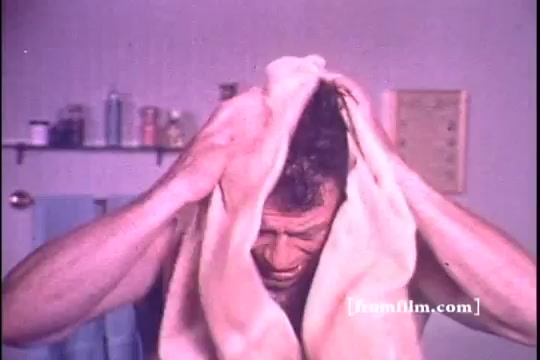 The still from the 1970s 'Schick Super II' commercial features a man in a bathroom, likely in the midst of grooming. He is using a towel to dry his hair, with a focused expression. The setting includes various toiletries and a simple, clean background, typical of that era's advertising aesthetics. The color tone reflects the period's film quality, giving it a nostalgic feel.