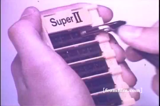 The still features a hand holding a Schick Super II razor cartridge holder, showcasing its design from the 1970s. The cartridge holder is labeled 'Super II' and contains several slots for razor cartridges. The advertisement emphasizes ease of use, reflecting the branding and marketing style of that era. The visual style indicates it's from a 16mm print, characteristic of vintage commercials.
