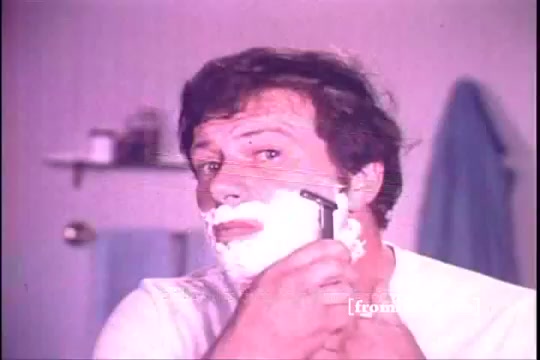 The still from the 'Schick Super II' commercial features a man in a bathroom, applying shaving cream to his face. He is using a razor while looking into the mirror. The scene captures the essence of personal grooming typical of the 1970s, with a color tone that reflects the era's aesthetic. The background includes bathroom items, emphasizing a domestic setting.
