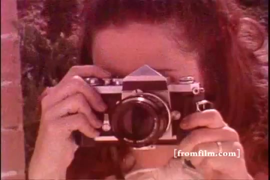 The still features a young woman with wavy hair, holding a vintage camera to her face, poised to take a photograph. The image has a warm, nostalgic tone typical of 1970s film, with soft focus and slightly faded colors. The background hints at outdoor surroundings, suggesting a casual and sunny moment.