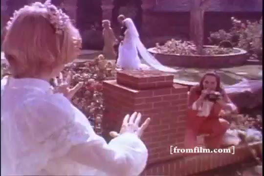 The still from the 1970s Bold Detergent commercial features a garden setting. A bride in a long white dress stands in the background, while a woman in a red outfit crouches in the foreground, seemingly taking a photo. The scene conveys a cheerful, celebratory atmosphere typical of domestic life and family moments. The color palette is warm, characteristic of the era's aesthetic.