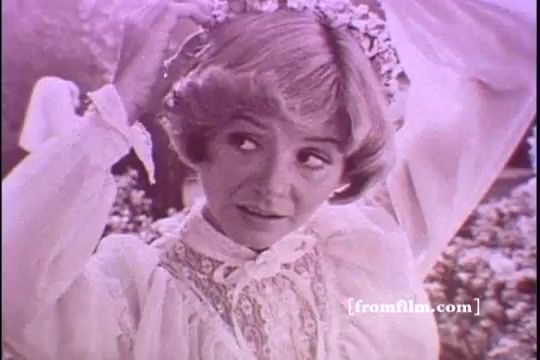 The still features a young woman with short, light-colored hair, adorned with a floral crown. She wears a vintage, frilly white dress and looks playfully at the camera with a slight smile. The background showcases greenery and blooms, giving a sunny, cheerful vibe typical of 1970s advertisements. The overall tone of the image has a soft, pastel quality, characteristic of the era's print style.