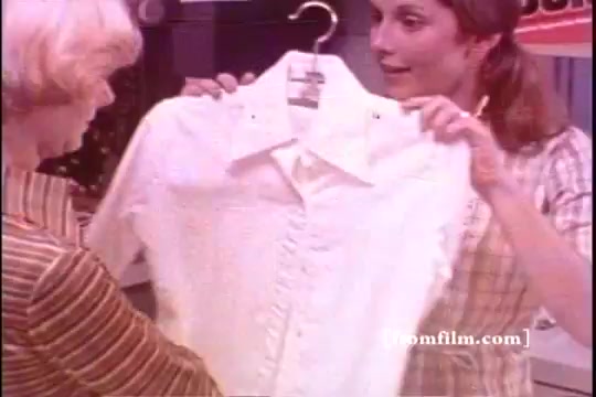 The still from 'Bold Detergent - 1970s' depicts two women in a store setting. One woman with light hair is showcasing a white shirt hanging on a hanger, while the other woman, wearing a striped blazer, appears to engage with interest. The scene illustrates a cheerful, consumer-driven moment typical of the era's advertising. The background features colorful signage hinting at the product's promotion.