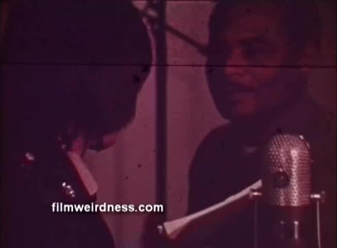 The still from the PSA 'Join the people who've joined the army' features two individuals engaged in a conversation. One person appears to be speaking into a microphone, likely in a journalistic context, while the other is listening attentively. The setting suggests a discussion about the army's opportunities in photo journalism, highlighting the training and skills acquired through the military. The image has a vintage look consistent with 16mm film, contributing to the overall nostalgic vibe of the recruiting message.