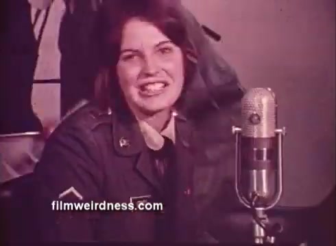 The still features a young woman in military uniform sitting in front of a microphone, smiling as she speaks. She emphasizes her curious nature and her choice of journalism as a career. The context is a U.S. Army recruiting public service announcement (PSA), promoting the Army's photojournalism course as a fast-tracked path to a journalistic career. The setting appears informal, likely in a studio or casual environment.