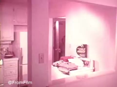 The still from 'Cold Power Clothes Detergent' features a domestic kitchen scene with a soft pink hue. In the background, the kitchen is partially visible, showcasing white cabinetry and appliances. A doorway leads into another room, hinting at a domestic setting where laundry or household activities may take place. In the foreground, there are stacks of folded clothes, possibly ready for washing. The overall atmosphere suggests a focus on cleanliness and the efficacy of the detergent.   .