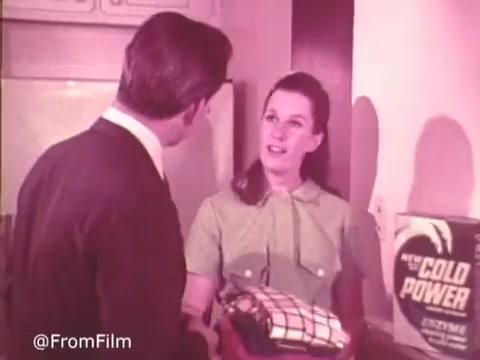 In this 16mm still from a Cold Power Clothes Detergent ad, a man in a suit converses with a woman wearing a light green dress. The woman is holding a folded laundry item while standing next to a prominently displayed box of Cold Power detergent. The warm, retro color palette adds a nostalgic feel to the scene, emphasizing domestic life and the product's appeal.   .