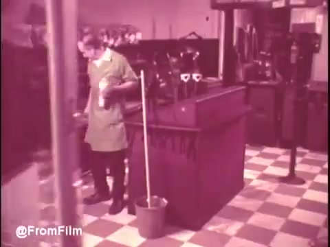 The still from the 'Ajax White Tornado' commercial features a woman in a green uniform, possibly cleaning, in a retro store setting with a checkered floor. The vintage aesthetic reflects the 1970s era, with household cleaning products likely in focus. The scene conveys a sense of everyday life and the promotion of Ajax's cleaning abilities.   .