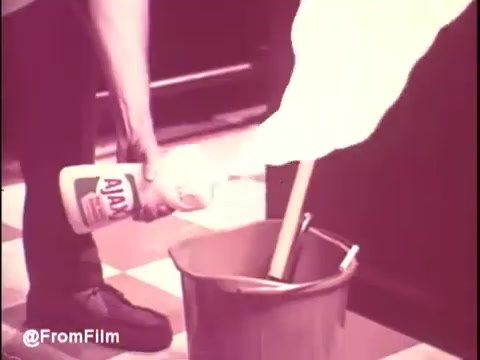 The still from the 'Ajax White Tornado' commercial features a person pouring Ajax cleaning solution into a bucket, likely preparing for cleaning. The scene is set against a classic checkered floor, with a focus on the action of mixing the cleaning product with water in the bucket. The colors may appear slightly muted or retro, typical of 1970s commercials. The commercial likely emphasizes the product's effectiveness in tackling tough cleaning tasks.   .