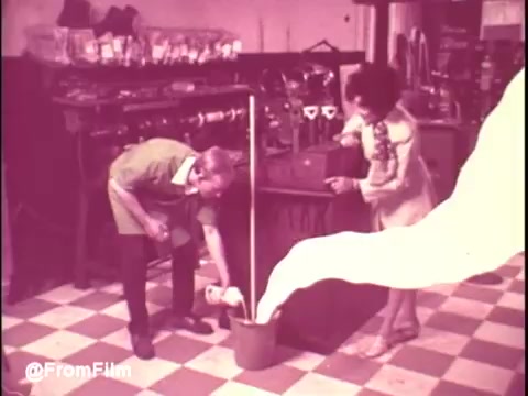 The still from the 'Ajax White Tornado' commercial features a young man in a green shirt pouring a cleaning product into a bucket while a woman watches with interest. The scene is set in a cluttered space, likely a store or kitchen, with various items and a checkered floor visible. The white product creates a swirling visual effect, emphasizing its cleaning power. The use of bright colors and playful expressions conveys the energetic vibe typical of 1970s commercials.   .