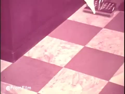 The still features a checkered floor, likely in a kitchen or living space, showcasing a cleaning scene typical of the 1970s. The color palette has a warm, faded quality, reflecting the vintage style of the era. A cleaning tool is visible, hinting at the promotional nature of the Ajax White Tornado product. The overall aesthetic captures the nostalgic vibe of classic commercials from that time.   .