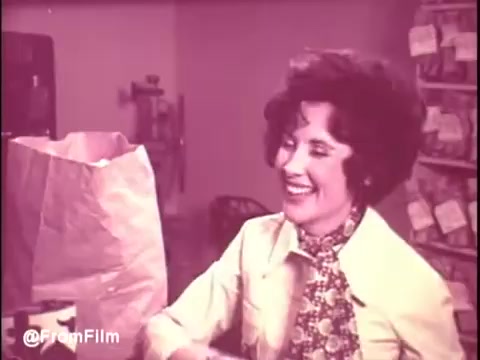 The still from the Ajax White Tornado commercial features a smiling woman with curly hair, dressed in a light-colored top and patterned scarf. She appears to be in a homey setting, surrounded by bags and domestic items. The overall tone is warm and cheerful, reflecting the inviting nature of 1970s advertising. The color palette has a vintage quality typical of 16mm film.   .