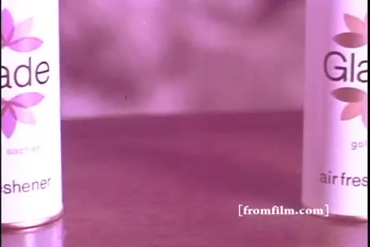 The still features two Glade air freshener cans placed on a surface, with a soft focus background creating a blurred effect. The cans are prominently labeled, showcasing the brand name and floral design. The color palette is warm, with a hint of vintage appeal typical of 1970s advertising aesthetics.