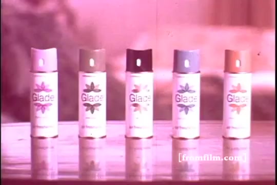 The still features five cans of Glade air freshener lined up on a table against a soft, muted background. Each can has a distinct color cap and displays the Glade logo along with floral motifs. The image has a gentle, warm tone typical of 1970s advertising, suggesting a focus on home ambience and freshness. The presentation looks cheerful and inviting, aimed at promoting various fragrance options.