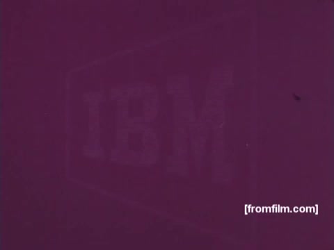 The still features a muted, purple backdrop with the faint outline of the IBM logo prominently displayed. This 16mm print captures a moment from 'IBM: Once Upon A Punched Card' (1964), highlighting the historical significance of punch cards in computing. The image evokes a sense of nostalgia, reflecting an era when such technology was revolutionary. Follow us on Twitter @FromFilm for more vintage insights!