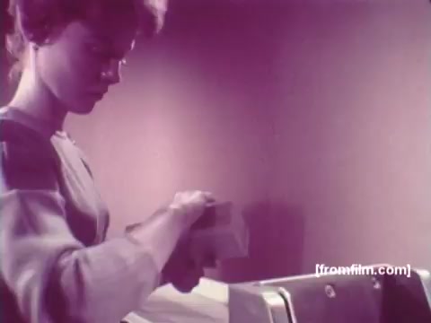 The still from 'IBM: Once Upon A Punched Card' (1964) captures a moment of early computing where a woman is handling a stack of punch cards. The setting is likely an office or laboratory, and the color palette leans towards muted tones, reflecting the vintage aesthetic of the time. Her focused expression suggests a task involving data entry or processing, embodying the pivotal role of punch cards in the computing era. This film serves as an exploration of the evolution of punch cards and their significance in the history of technology.