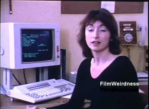 The still depicts a woman speaking in front of a desk with an IBM System 36 computer. The monitor displays text, suggesting a demonstration or explanation related to the system. The setting appears to be an office, with a vintage phone visible in the background. The overall tone is professional and showcases early computing technology from 1984.