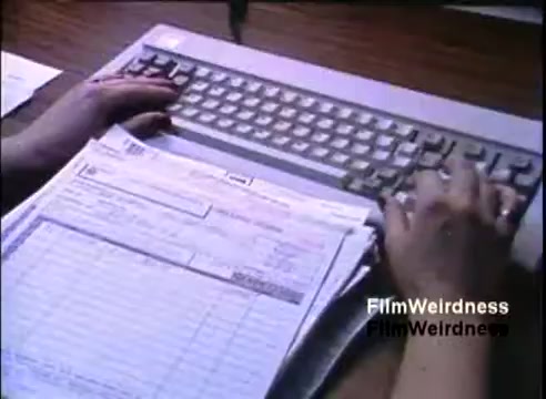 The still features a close-up of a person’s hands typing on a keyboard. In front of them is a printed document, possibly a form or report, indicating administrative or business tasks. The scene emphasizes the use of the IBM System 36, showcasing the technology's role in office work during the 1980s. The layout reflects a classic workstation setup of that era.
