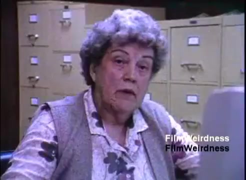 The still features an older woman sitting at a desk, looking straight at the camera. Behind her is a filing system, suggesting an office environment. She appears to be engaged in a discussion, likely about the IBM System 36, given the context of the film. The setting exudes a typical 1980s office atmosphere.