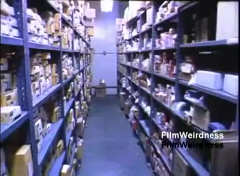 The still depicts a narrow warehouse aisle lined with metal shelves filled with boxes and products, likely from the 1980s. The lighting is fluorescent, casting a stark light on the items, which include various technology and hardware components associated with the IBM System 36. The scene conveys a sense of inventory and stock management typical in a sales or distribution setting for tech products from that era.
