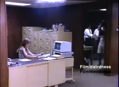 The still from the IBM System 36 sales film (1984) depicts an office scene. On the left, a woman is seated at a desk, focused on paperwork, with a small computer nearby. In the background, two individuals are entering the office, suggesting a collaborative or interactive work environment. Filing cabinets line the wall, hinting at a traditional office setup typical of the era. The lighting is somewhat subdued, contributing to the retro ambiance of the scene.