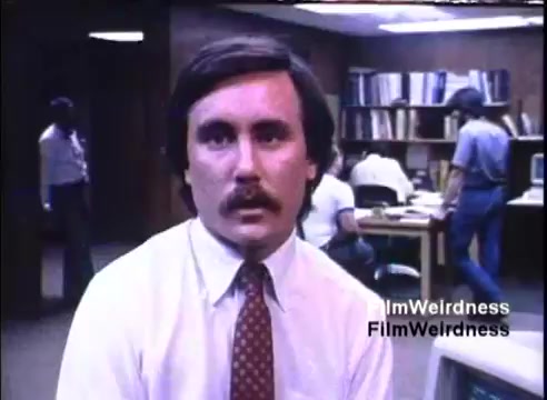 The still features a man in a white shirt and patterned tie, seated in an office environment. He appears to be speaking directly to the camera, with a serious expression. In the background, several people are engaged in work at desks, indicative of a busy corporate setting. The lighting is subdued, typical of office spaces from the 1980s, creating a professional atmosphere.