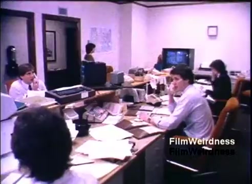 The still depicts an office environment in 1984, featuring several individuals engaged in work activities. There are multiple desks cluttered with papers and office supplies. A couple of people are on phones, while a television screen is visible in the background, showcasing a typical workplace setup of that era. The overall atmosphere suggests a busy, professional setting focused on productivity, likely related to the promotion of IBM's System 36.