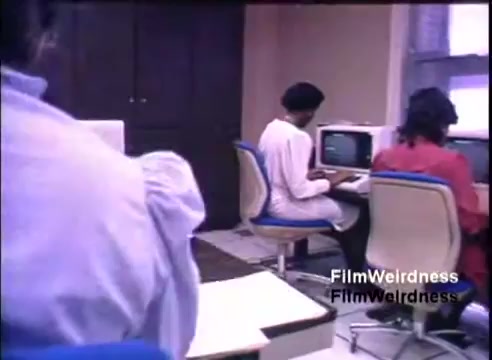 The still shows a small office setting from the 1984 IBM System 36 sales film. It features three individuals seated at computer workstations, focused on their screens. One person is dressed in white, and another in red, while a third, partially visible, wears a blue top. The environment appears professional, with wooden cabinets in the background and small computer monitors in front of the users. The overall tone suggests a contemporary office scene highlighting the use of technology in the workplace.