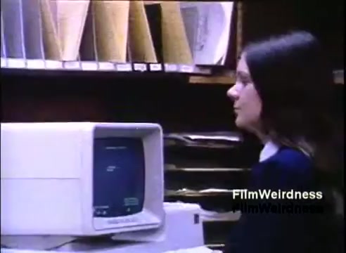 The still shows a young woman seated at a desk in an office setting, interacting with an IBM System 36 computer. The monitor displays text, suggesting a business application or data entry. Behind her, there are file folders neatly organized, indicating a focus on administrative tasks. The overall atmosphere reflects a typical work environment from the 1980s, highlighting the computer's role in office productivity.