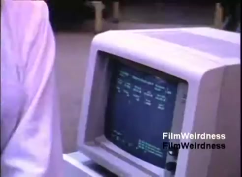 The still depicts an IBM System 36 computer setup from the 1984 sales film. The screen shows text-based data or commands, typical of early computing interfaces. A person stands partially in the frame, wearing a light-colored garment, indicating a formal or business context. The overall scene conveys a professional environment where technology is being demonstrated.