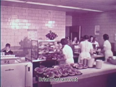 The still from the 'USDA School Lunch Program PSA - 1967' depicts a bustling school cafeteria environment. In the foreground, trays of food are prominently displayed, showcasing items likely served to students. Behind the counter, cafeteria workers in white uniforms are busy preparing and serving meals. A child is seated at the counter, observing the activity. The setting features tiled walls, typical of school cafeterias of that era, highlighting the focus on nutritious meals for children.