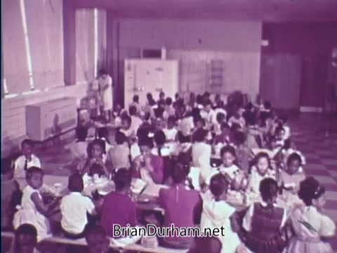 The still from the 1967 USDA School Lunch Program PSA depicts a school cafeteria scene with a large group of children seated at tables enjoying their meals. The setting is spacious and well-lit, featuring a variety of young students, predominantly in colorful clothing. In the background, a staff member appears to be supervising or assisting the children. The overall atmosphere emphasizes the communal experience of school lunches.