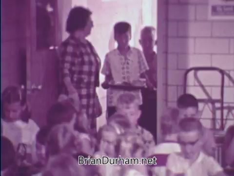 The still from the 1967 USDA School Lunch Program PSA shows a group of children seated at tables in a cafeteria. A young boy, carrying a tray, enters through a doorway alongside an adult woman in a checkered dress and another adult. The atmosphere suggests a typical school lunch setting, highlighting the program's focus on providing meals to children. The image captures a moment of community and nutrition in an educational environment.